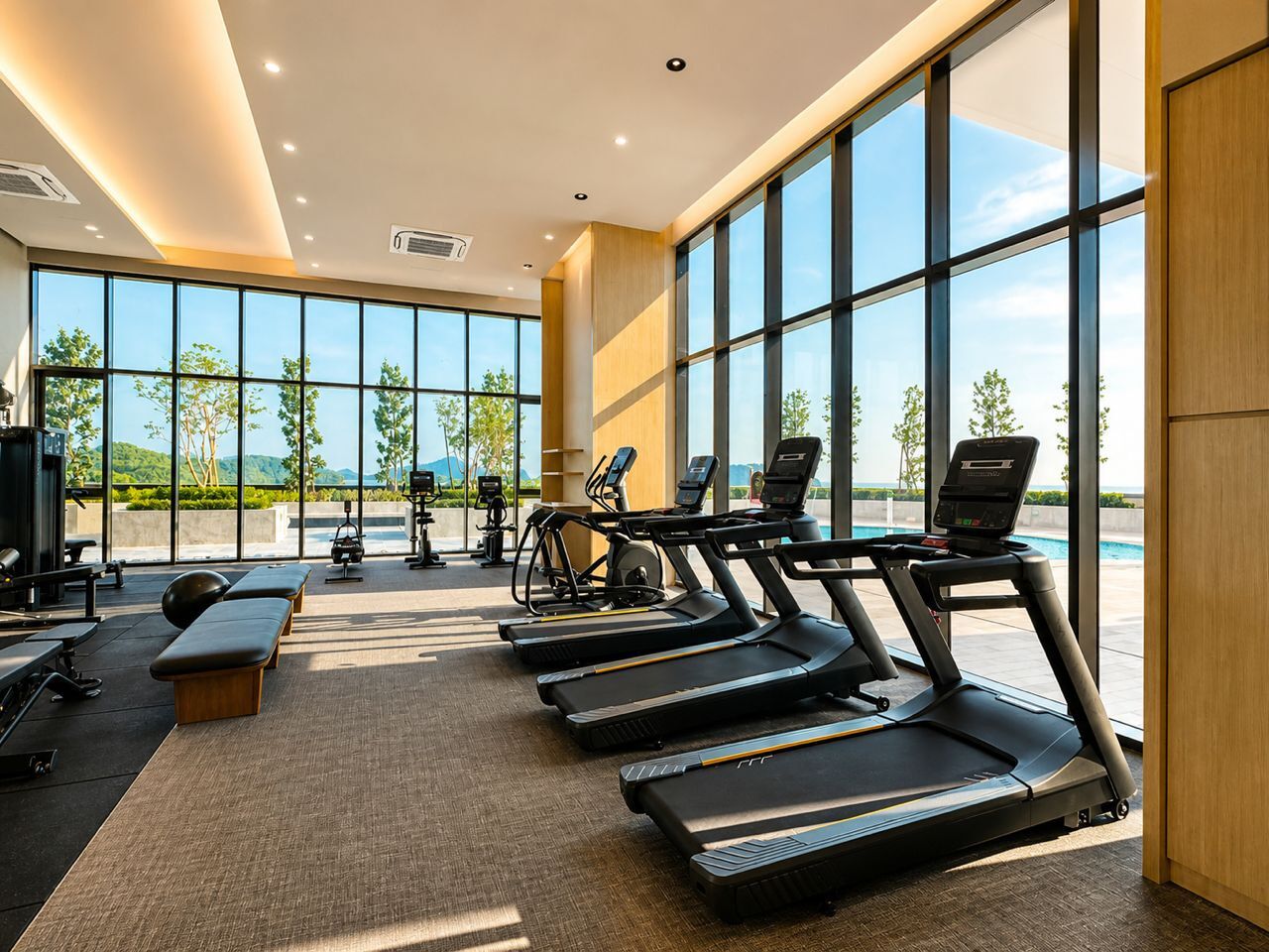 Ett gym med löpband, motionscyklar och träningsbänkar, stora fönster med utsikt över grönområden och en sjö eller ett annat vattendrag.