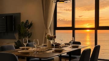 A dining area with a table set for four, large windows with a sunset view, and a plant on the left.