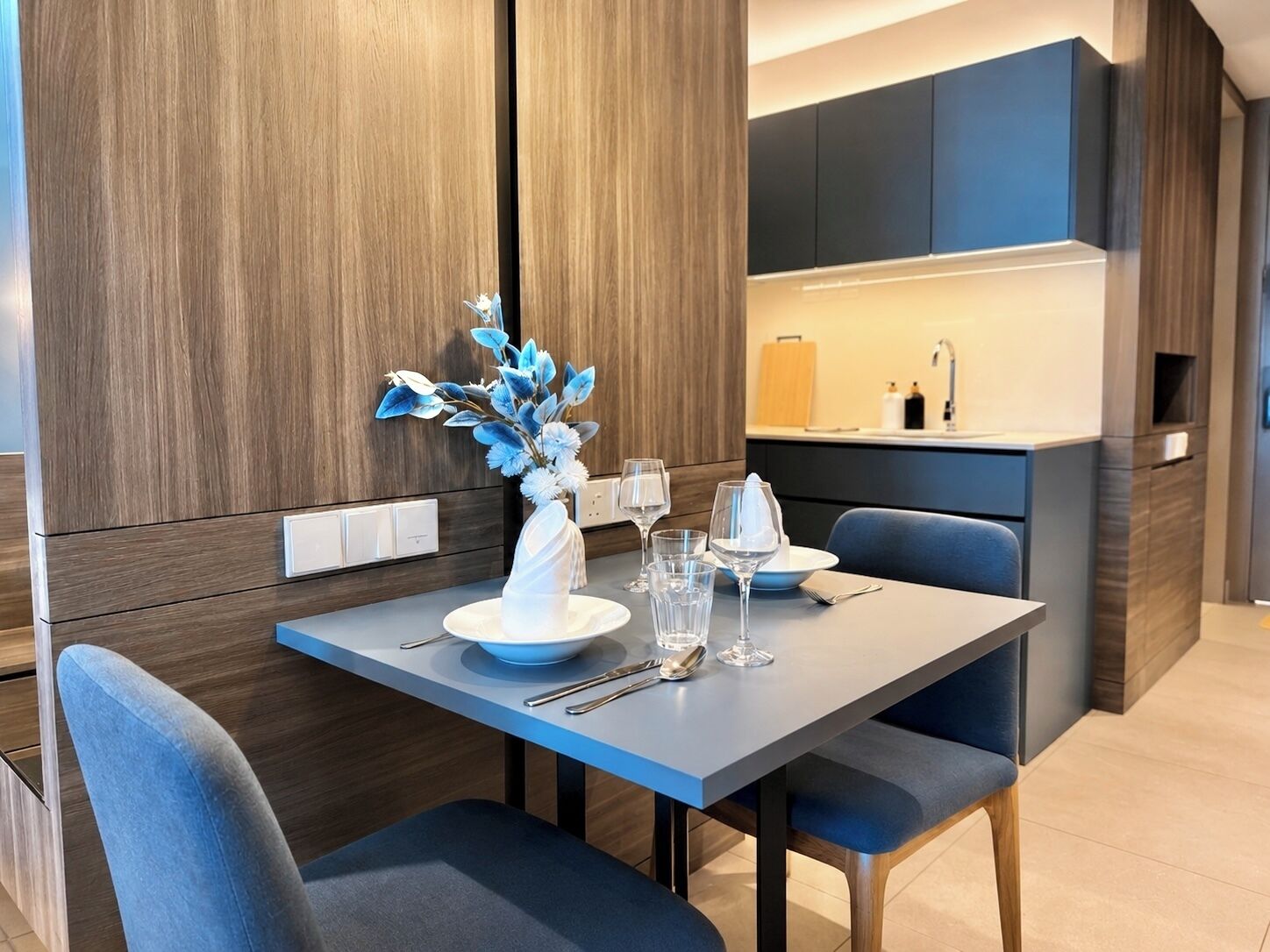 A modern dining area with a table set for two, a vase with blue flowers, and a kitchen in the background.