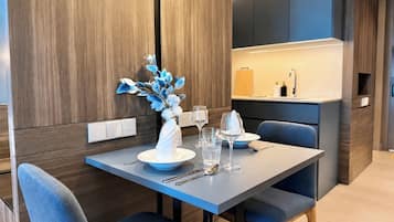 A modern dining area with a table set for two, a vase with blue flowers, and a kitchen in the background.