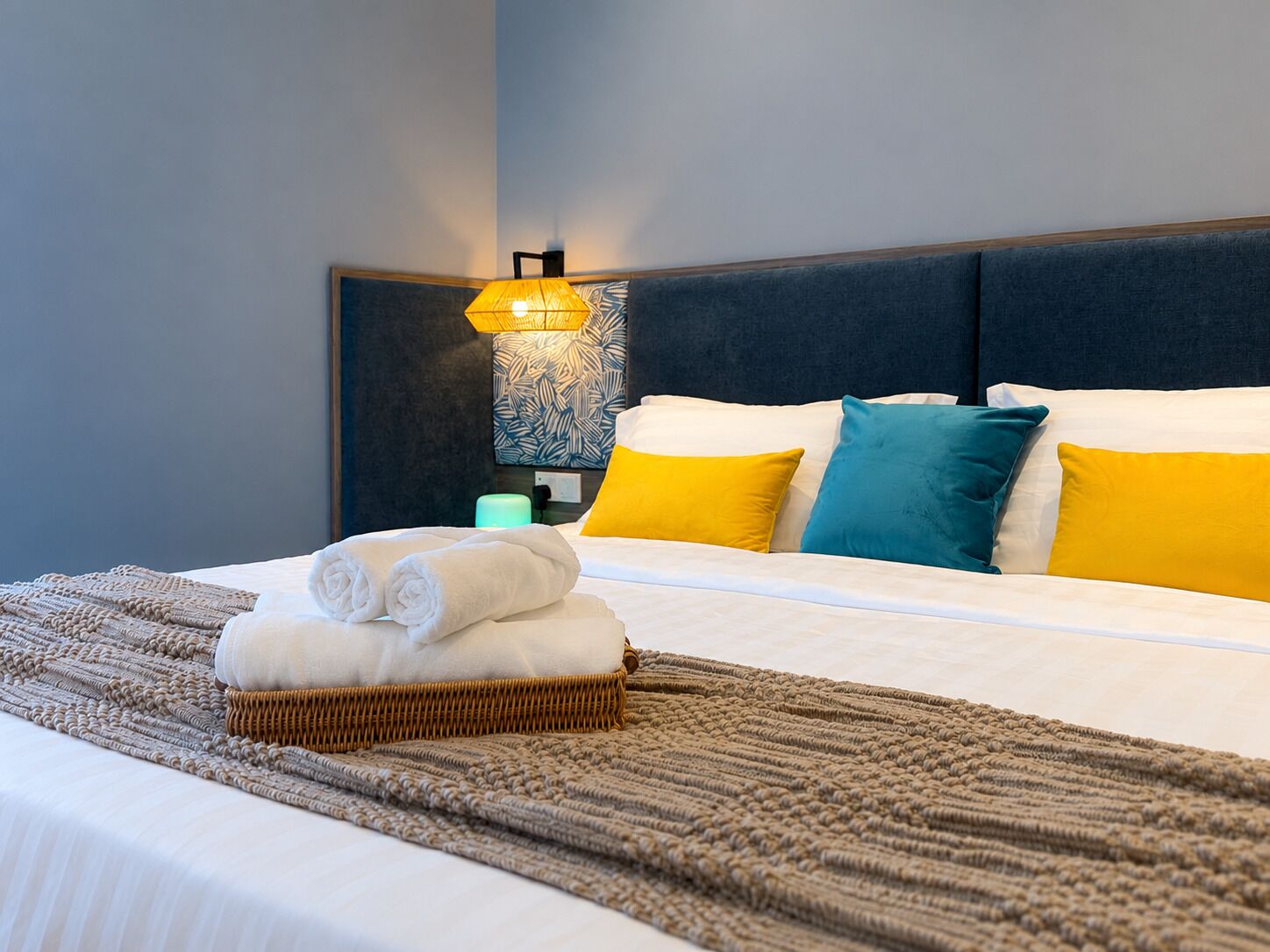 A neatly made bed with white linens, a woven basket with folded towels, and a wall-mounted lamp.