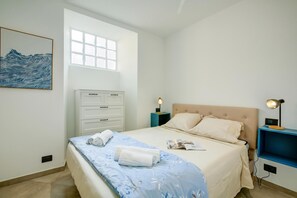 Ein Schlafzimmer mit Bett, Nachttisch, Kommode, Fenster und einem Wandbild.