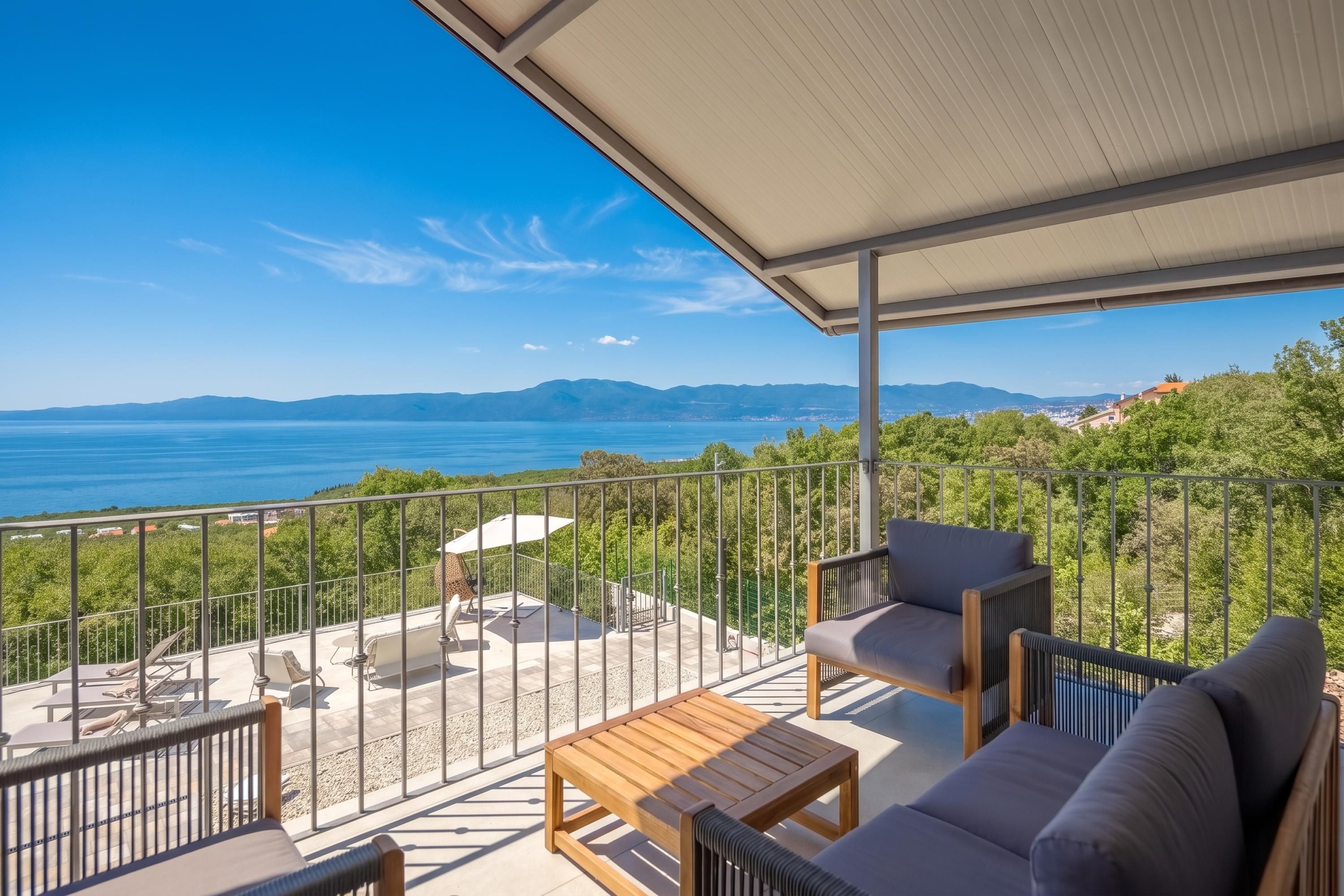 A balcony with a view of the sea and mountains, furnished with a sofa, chairs, and a table.