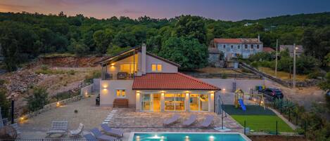 A two-story house with a swimming pool, outdoor seating, and a view of the surrounding landscape.