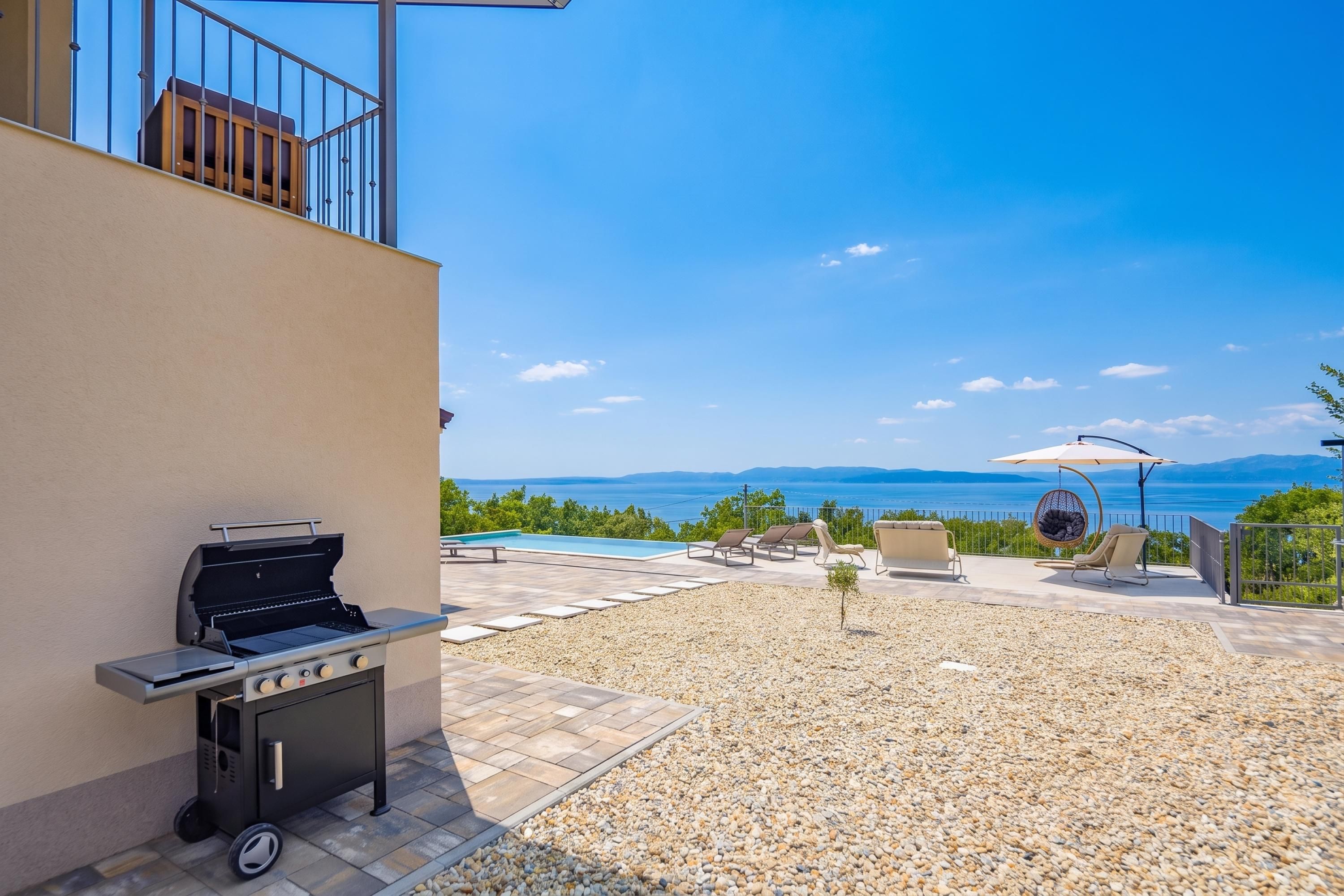 A patio with a grill, seating, and a pool overlooking a scenic view.