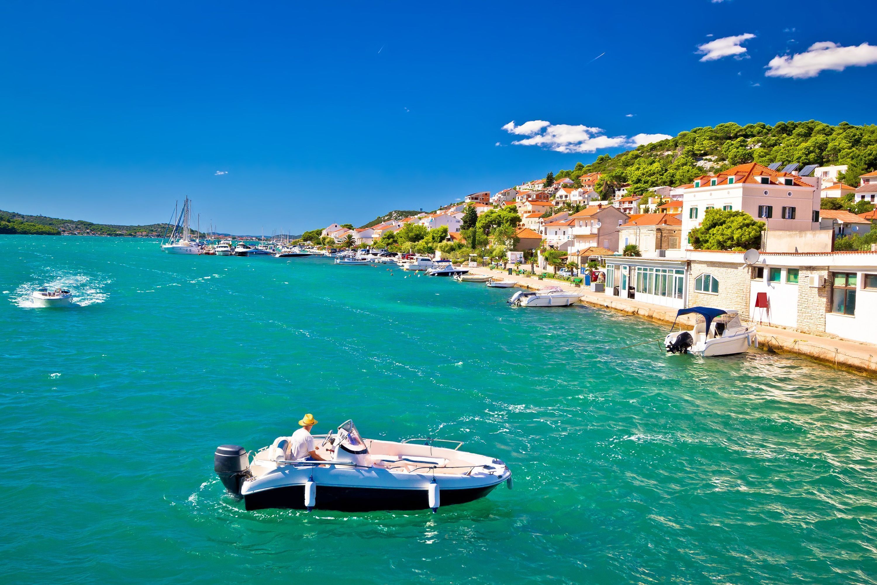 Une ville côtière où des bateaux sont amarrés le long du port, des bâtiments aux toits rouges et un ciel bleu azur.