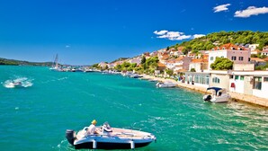 Uma vila costeira com barcos atracados na orla, edifícios com telhados vermelhos e um céu azul límpido.
