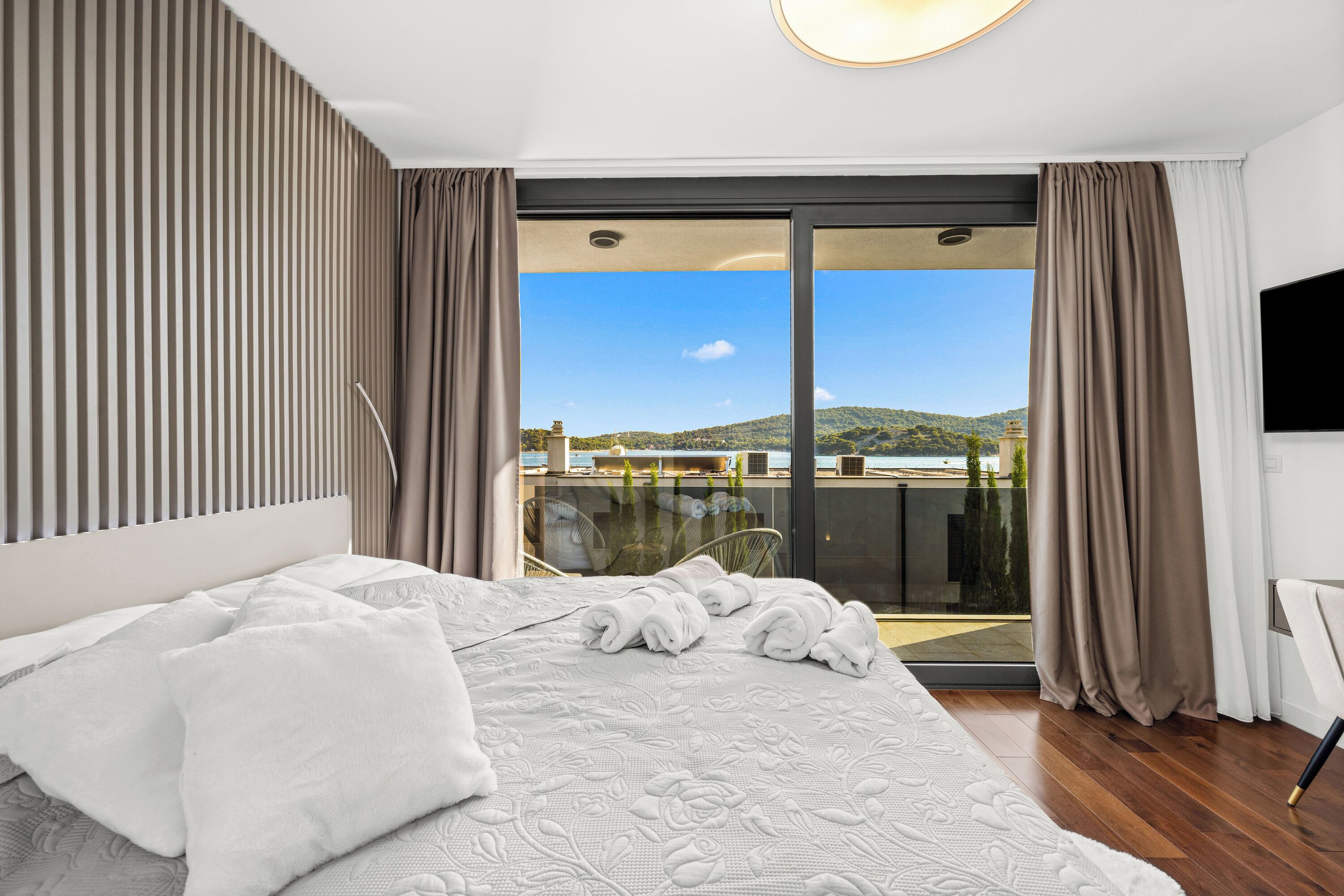 A modern bedroom with a large bed, a view of a balcony, and a cityscape in the background.