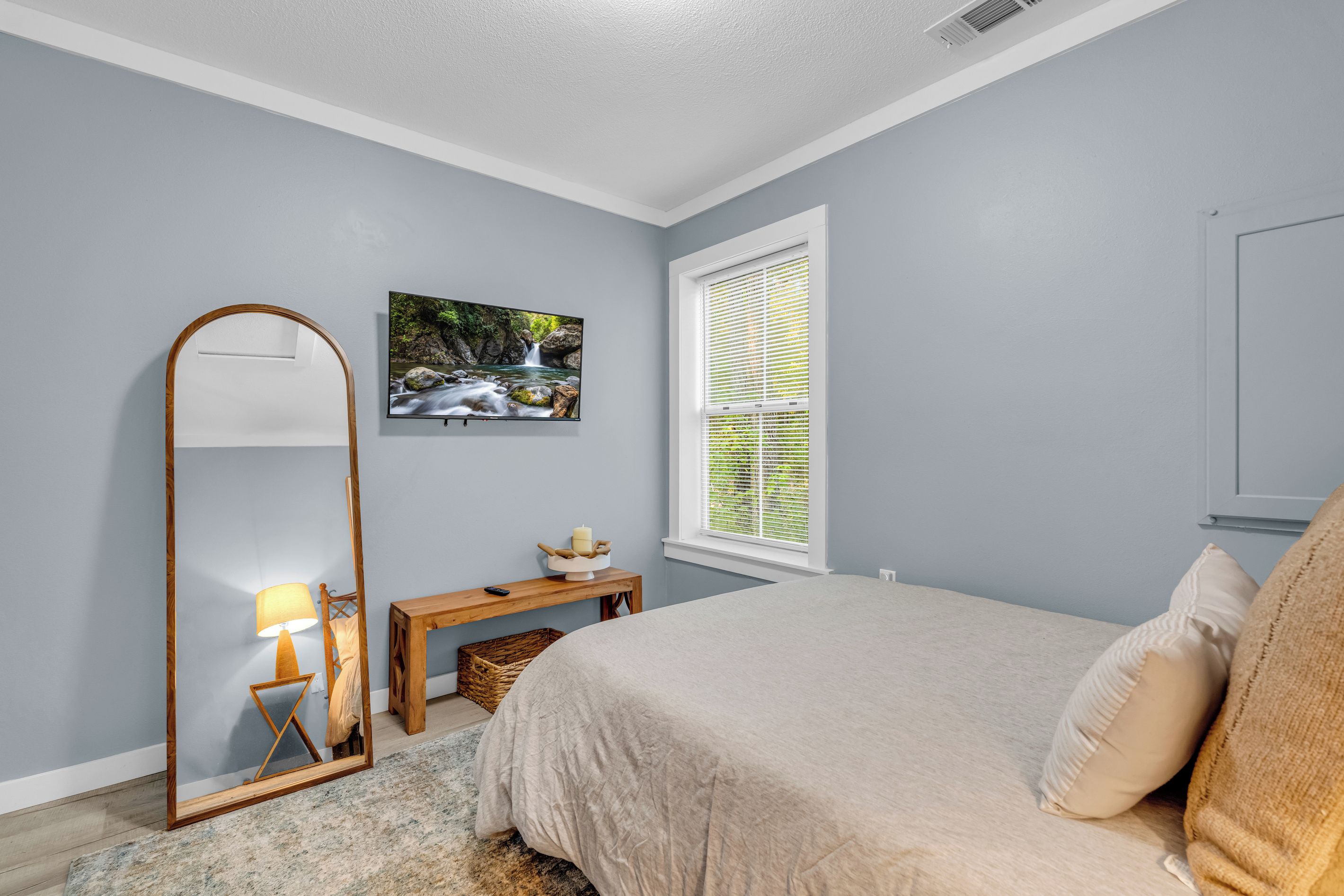A bedroom with a bed, a mirror, a bedside table with a lamp, a picture on the wall, and a window with blinds.