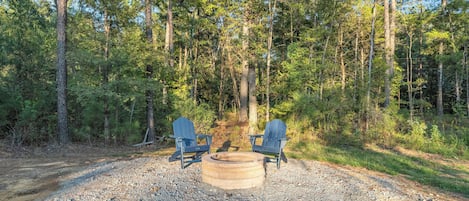 A forest campsite with two Adirondack chairs and a fire pit.
