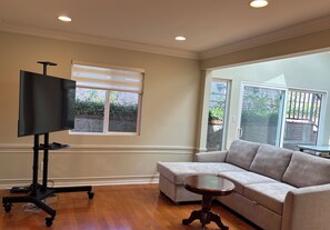 A living room with a flat-screen TV on a stand, a grey sofa, a small round table, and a window with blinds. - Elegant, peaceful LA home, 5 min from UCLA (Los Angeles)