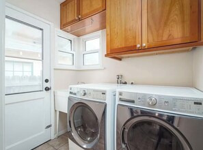 A laundry room with two front-loading washing machines, wooden cabinets, a white sink, and a window with blinds. - Elegant, peaceful LA home, 5 min from UCLA (Los Angeles)