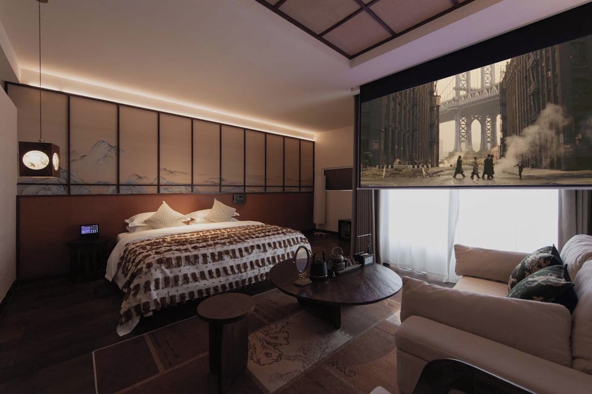 A modern hotel room with a large bed, a seating area, a television, and a view of a cityscape.