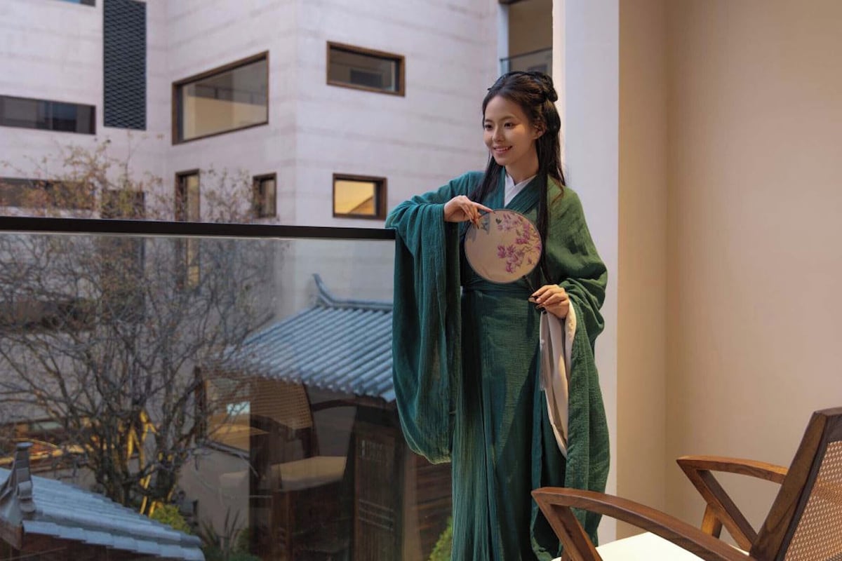 A person in traditional attire holding a fan, standing by a window overlooking a traditional building.