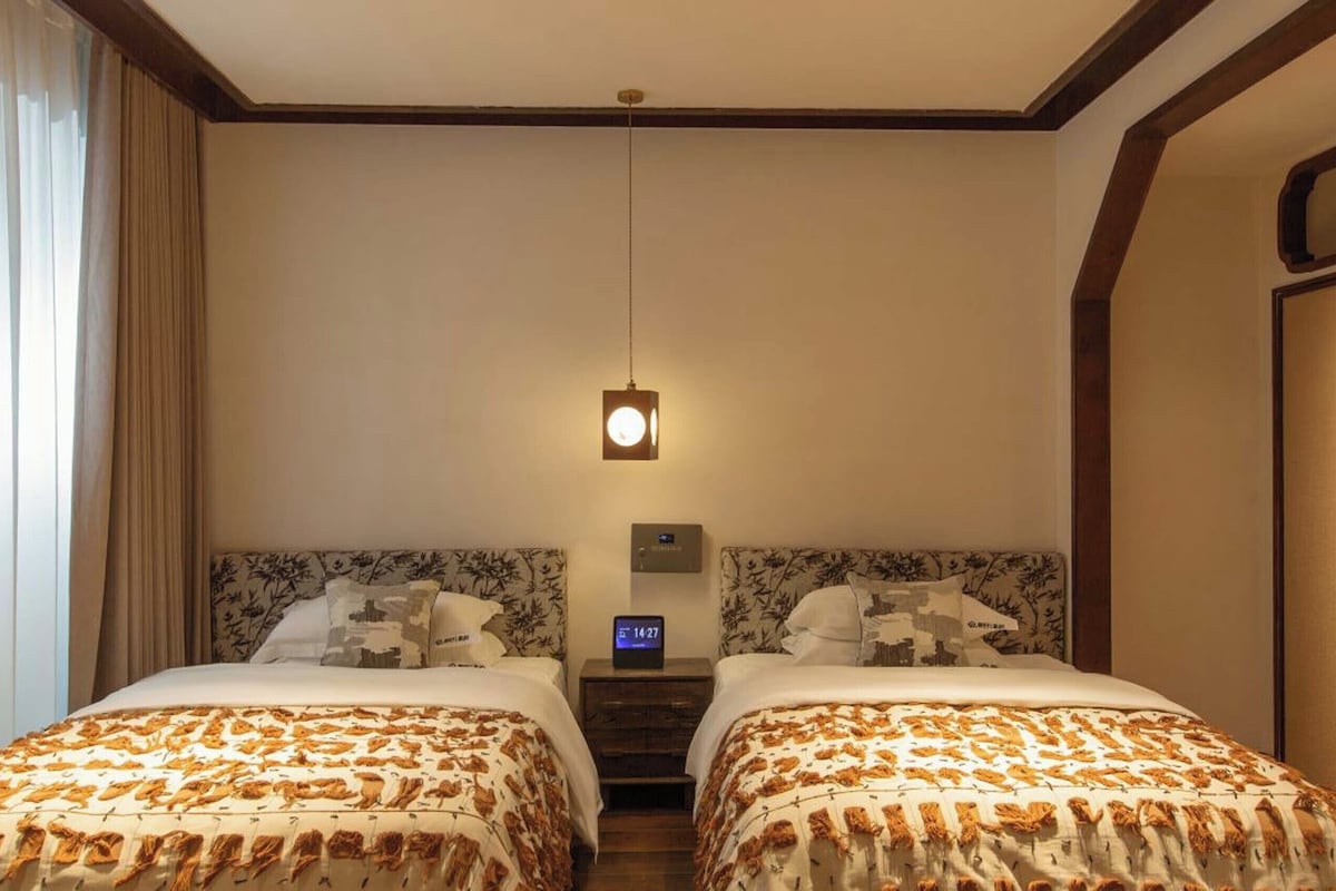 A hotel room with two beds, a nightstand with a clock, and a window with curtains.
