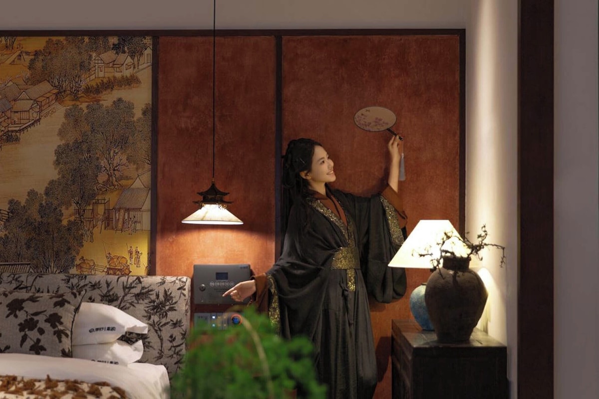 A room with a traditional Chinese painting, a bed with floral bedding, a desk with a lamp, and a person in traditional attire.