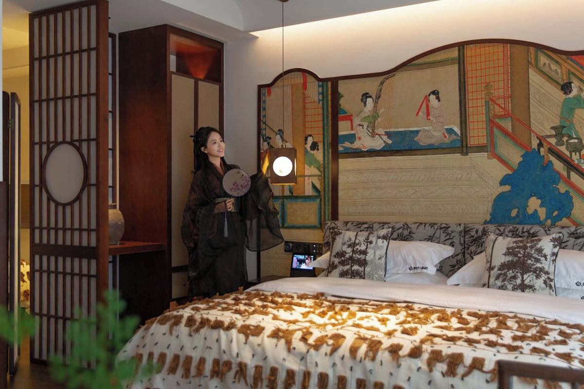 A traditional room with a bed, a wall with a painting, and a person holding a fan.