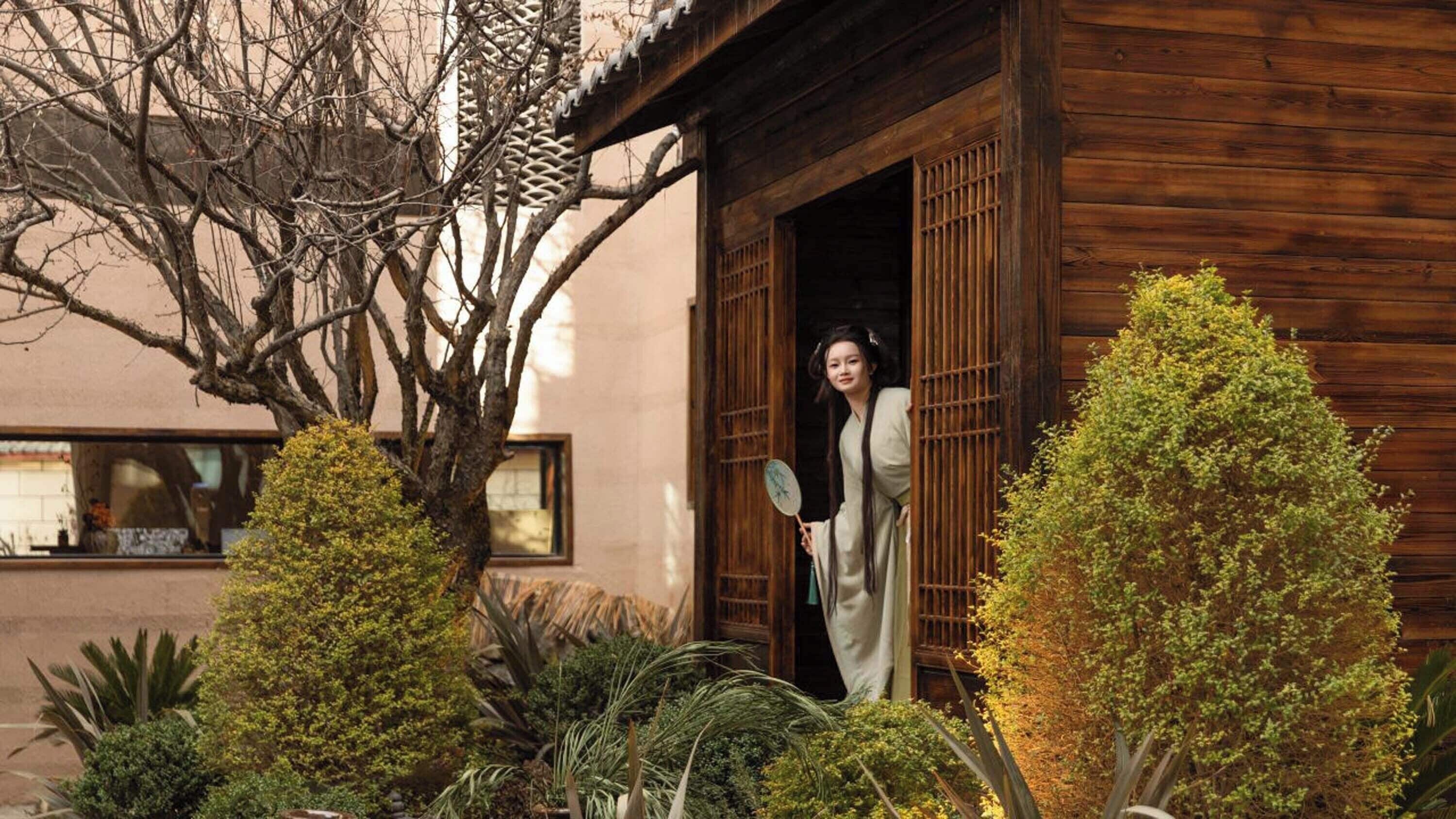 A traditional wooden building with a person standing at the entrance, surrounded by well-maintained greenery.