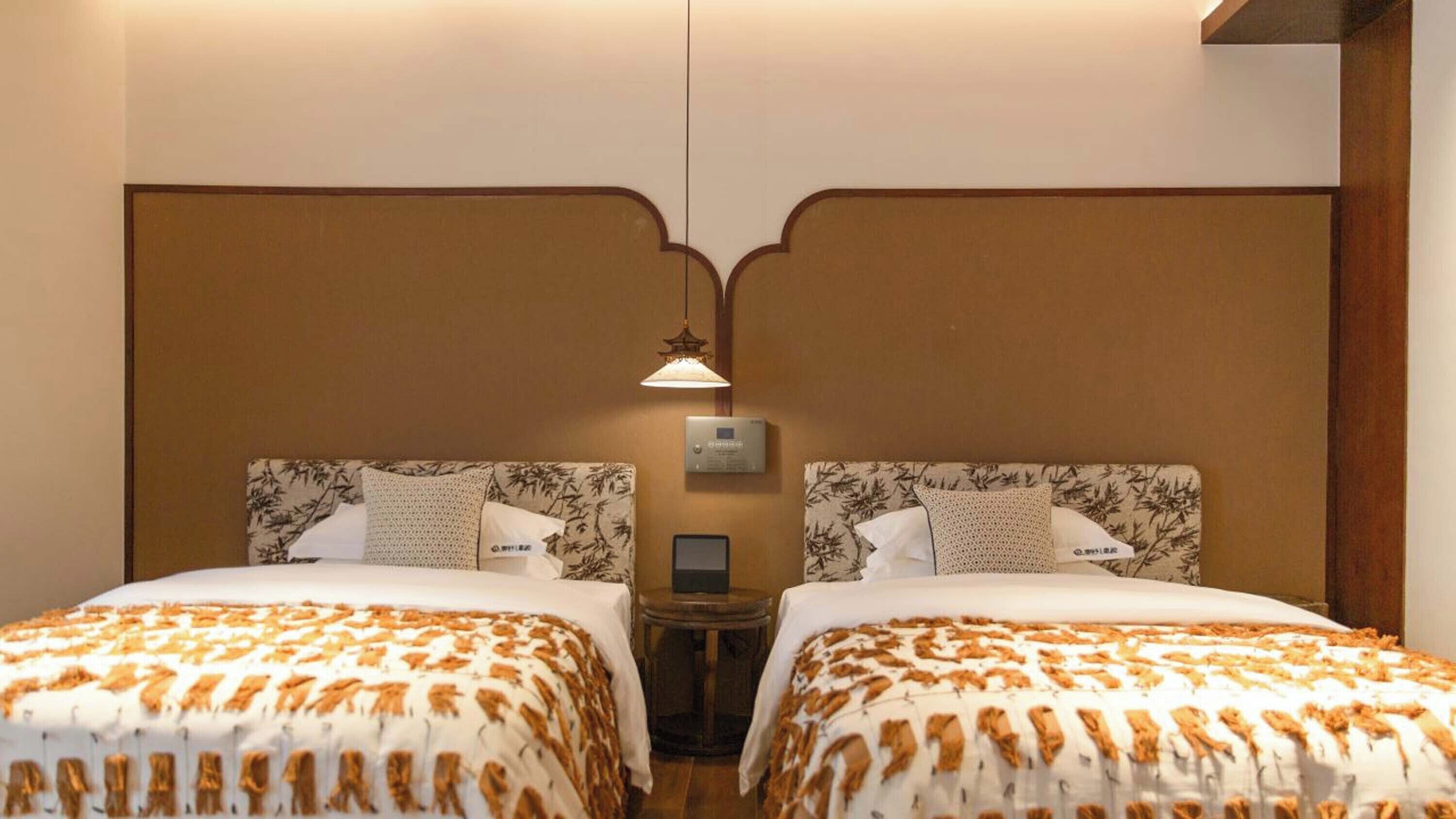 Une chambre d’hôtel avec deux lits, chacun doté d’une tête de lit décorée, d’une table de chevet et d’un panneau de commande fixé au mur.