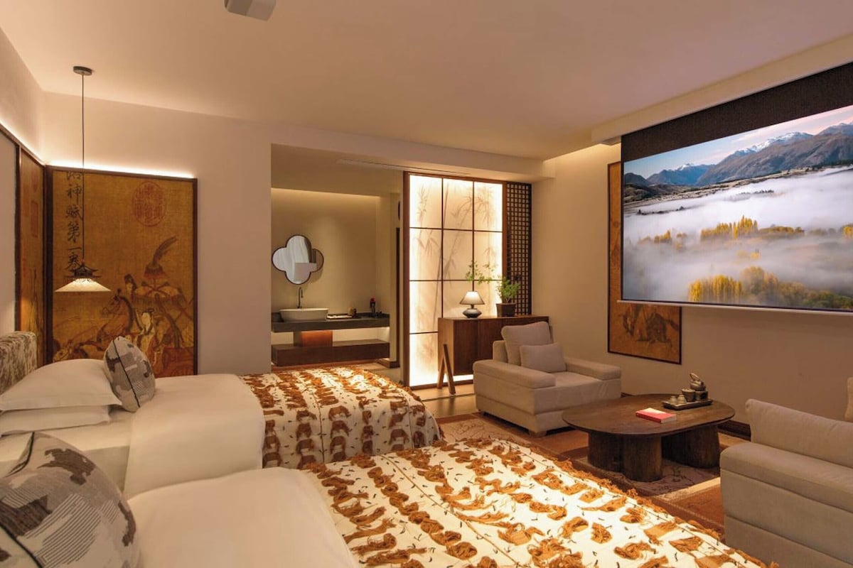 A modern hotel room with a large bed, two sofas, a coffee table, and a large window with a view of a mountainous landscape.