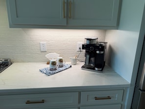 A kitchen with a coffee maker, two mugs, a white countertop, and a white brick wall.