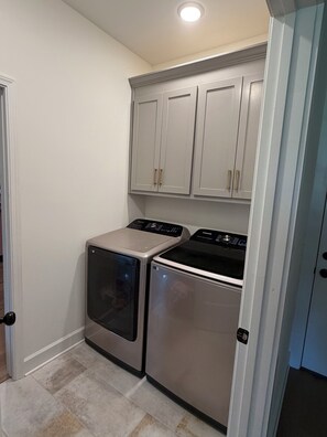 Un cuarto de lavandería con dos electrodomésticos de acero inoxidable, gabinetes blancos y piso de baldosas.