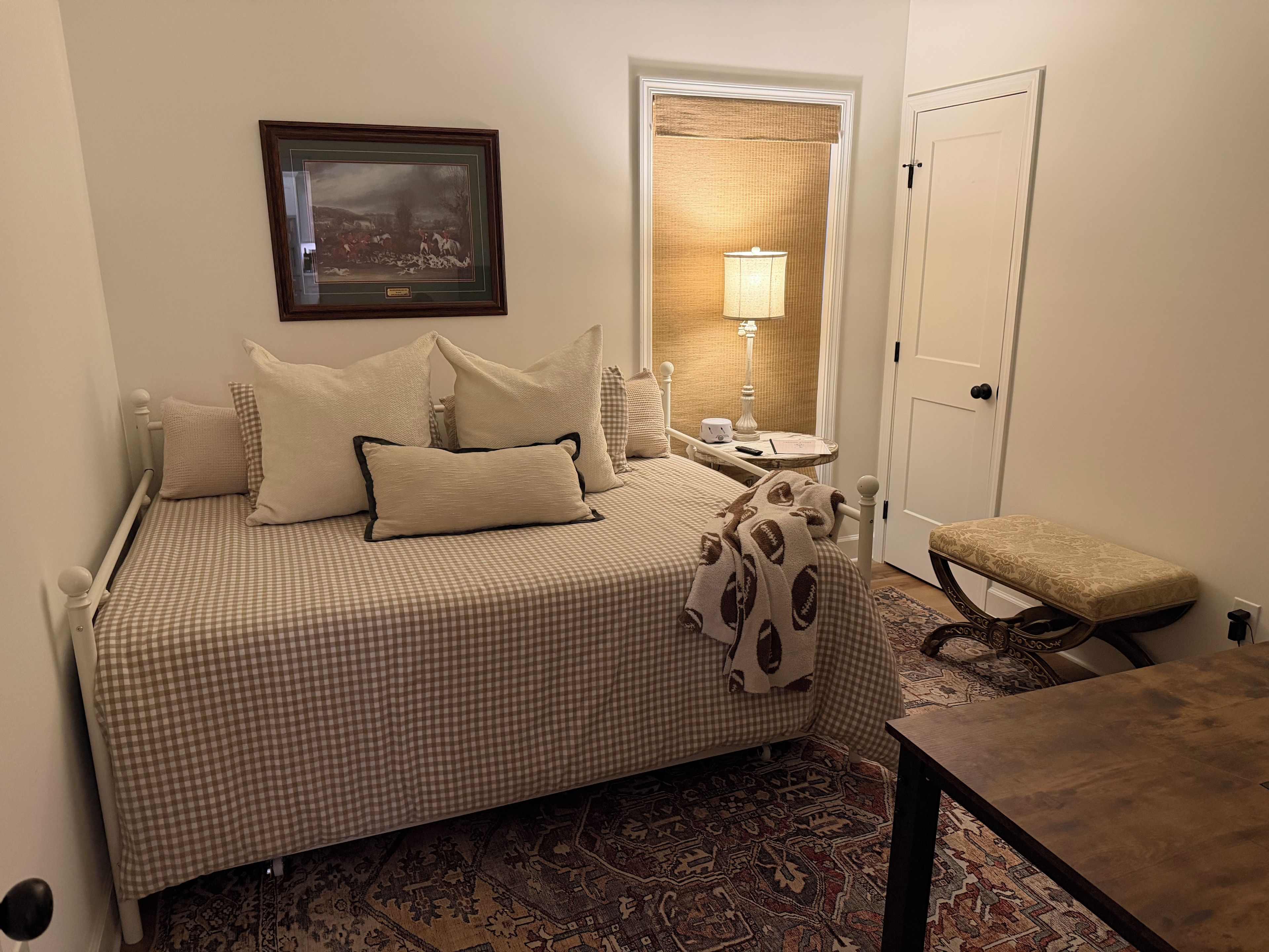 A neatly arranged bedroom with a checkered bedspread, a bedside lamp, a framed picture on the wall, and a bench.