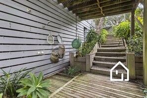 Un escalier en bois mène à une maison à l’extérieur blanc, entourée de verdure et d’un petit jardin.