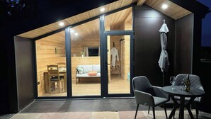 Eine moderne, hell erleuchtete Holzblockhütte mit Glastür, die zu einem Innenraum führt, der einen Essbereich, eine Couch und einen kleinen Tisch mit Stühlen beinhaltet.