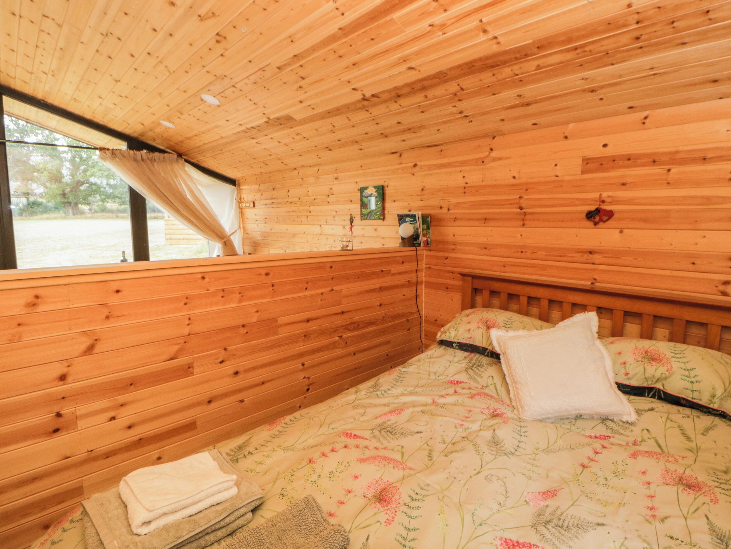 Ein Zimmer einer Holzhütte mit einem Bett, einem Fenster mit Vorhängen und einem kleinen Tisch mit einer Lampe.