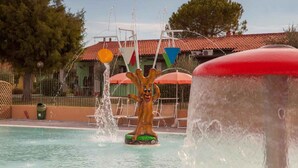Een waterpartij met een boomachtige sculptuur in een zwembadgedeelte, omringd door ligbedden en een gebouw met een rode pannendak.