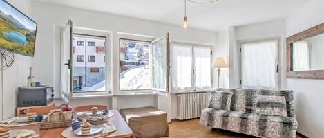 A cozy living room with a wooden floor, a sofa, a dining table with chairs, and a television displaying a snowy landscape.
