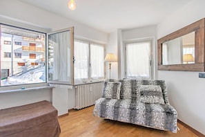 Ein Zimmer mit einer Couch, einem Fenster mit Blick auf ein Gebäude, einem Heizkörper und einem Spiegel an der Wand.