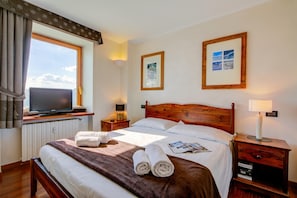 Una habitación de hotel con una cama, un televisor en un mueble bajo, una mesita de noche, una lámpara y un ventanal con cortinas.