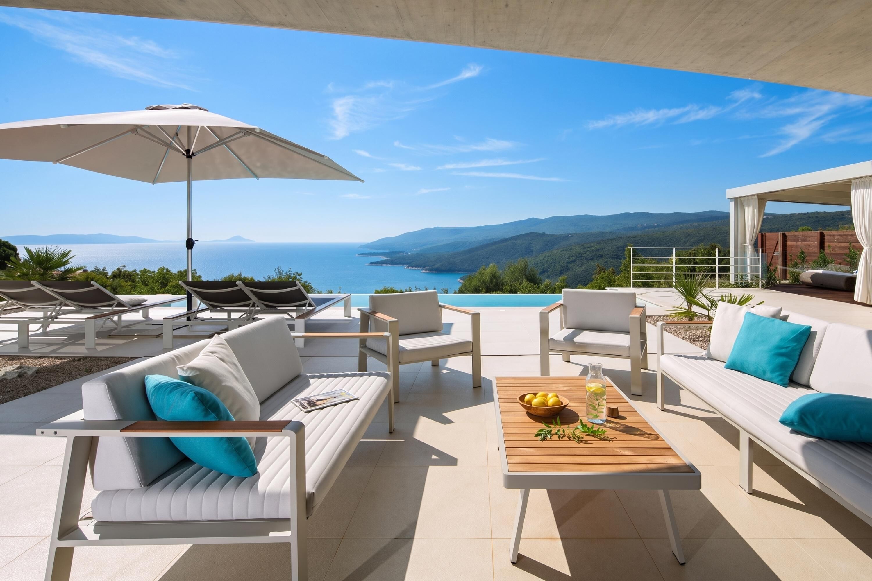 A terrace with white furniture, a wooden table with a bowl of lemons, and a view of the sea and mountains.