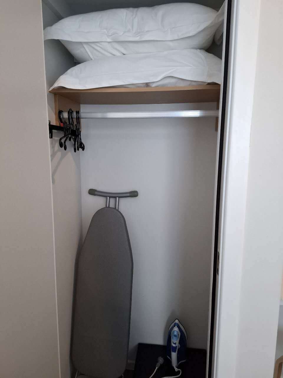 Ein Schrank mit Kleiderhaken, einem Regal voller gefalteter weißer Wäsche und einem Bügelbrett mit Bügeleisen.