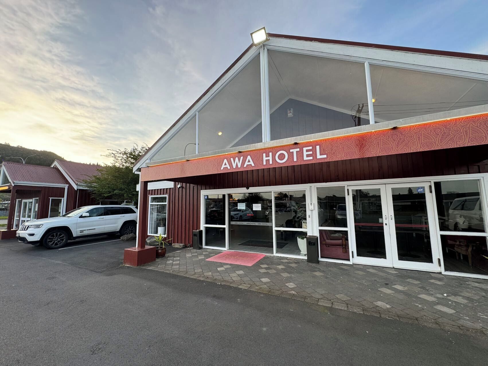 Een hotel met een grote glazen gevel, een rode luifel en een witte auto geparkeerd ervoor.