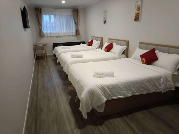 A hotel room with four beds arranged in a row, a flat-screen TV mounted on the wall, a window with curtains, and a small bedside table.