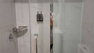A glass-enclosed shower stall with a handheld showerhead, a towel rack with a white towel, and a soap dispenser on the wall.