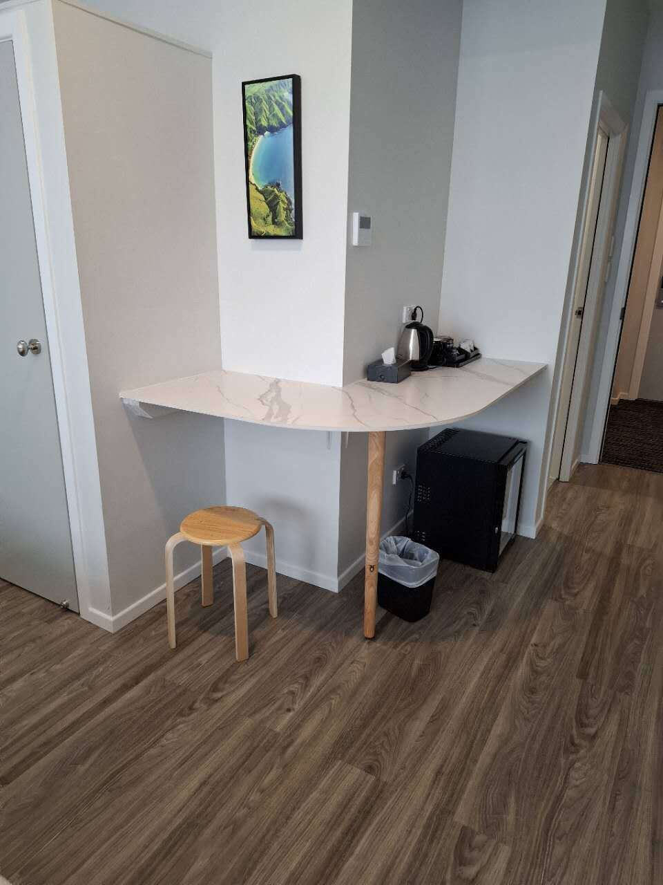 A compact room with a white marble desk, a small wooden stool, a black trash bin, and a black storage unit.