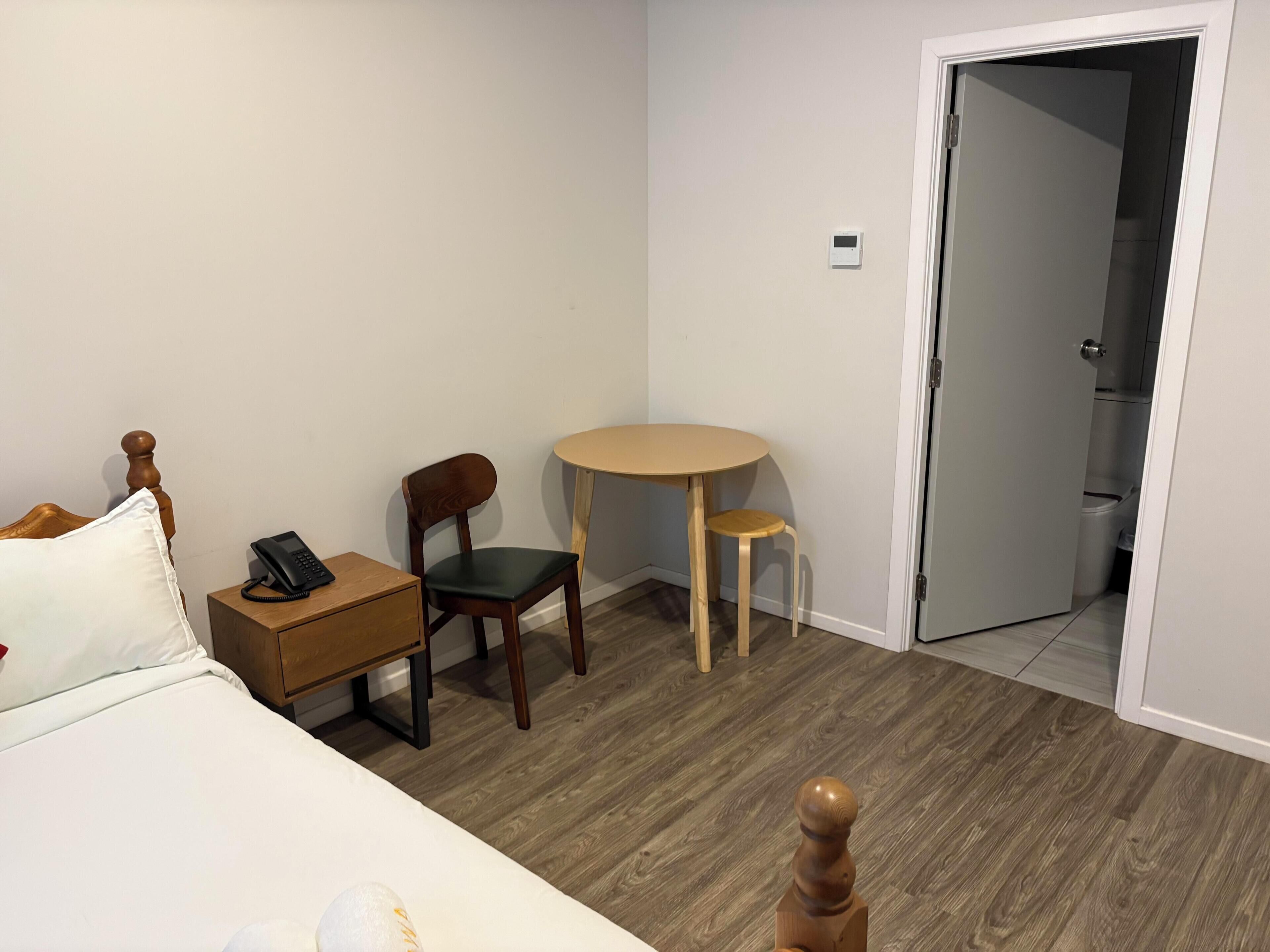 Habitación de hotel con cama, mesita de noche con teléfono, una silla, una mesita pequeña y una puerta que da al baño.