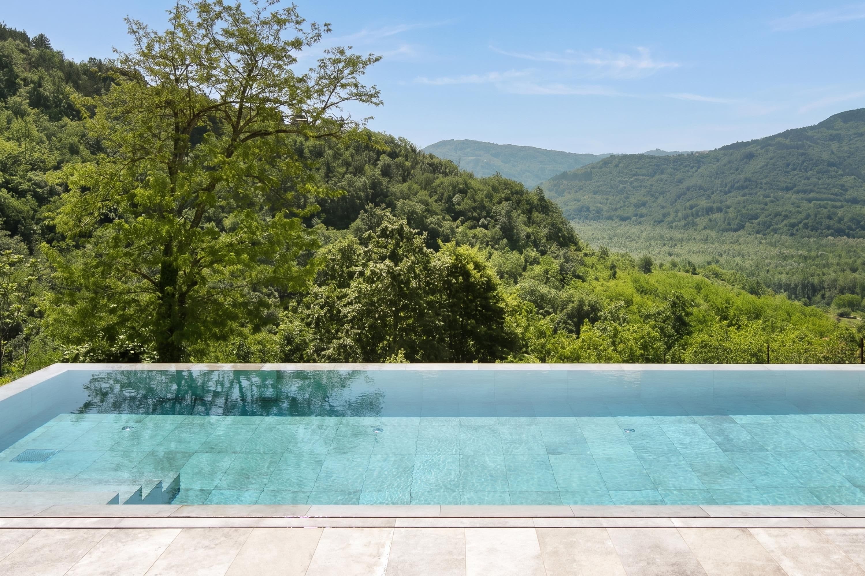 Uma piscina infinita retangular com água azul cristalina, rodeada por um deck de pedra, com vista para uma floresta verdejante e colinas.