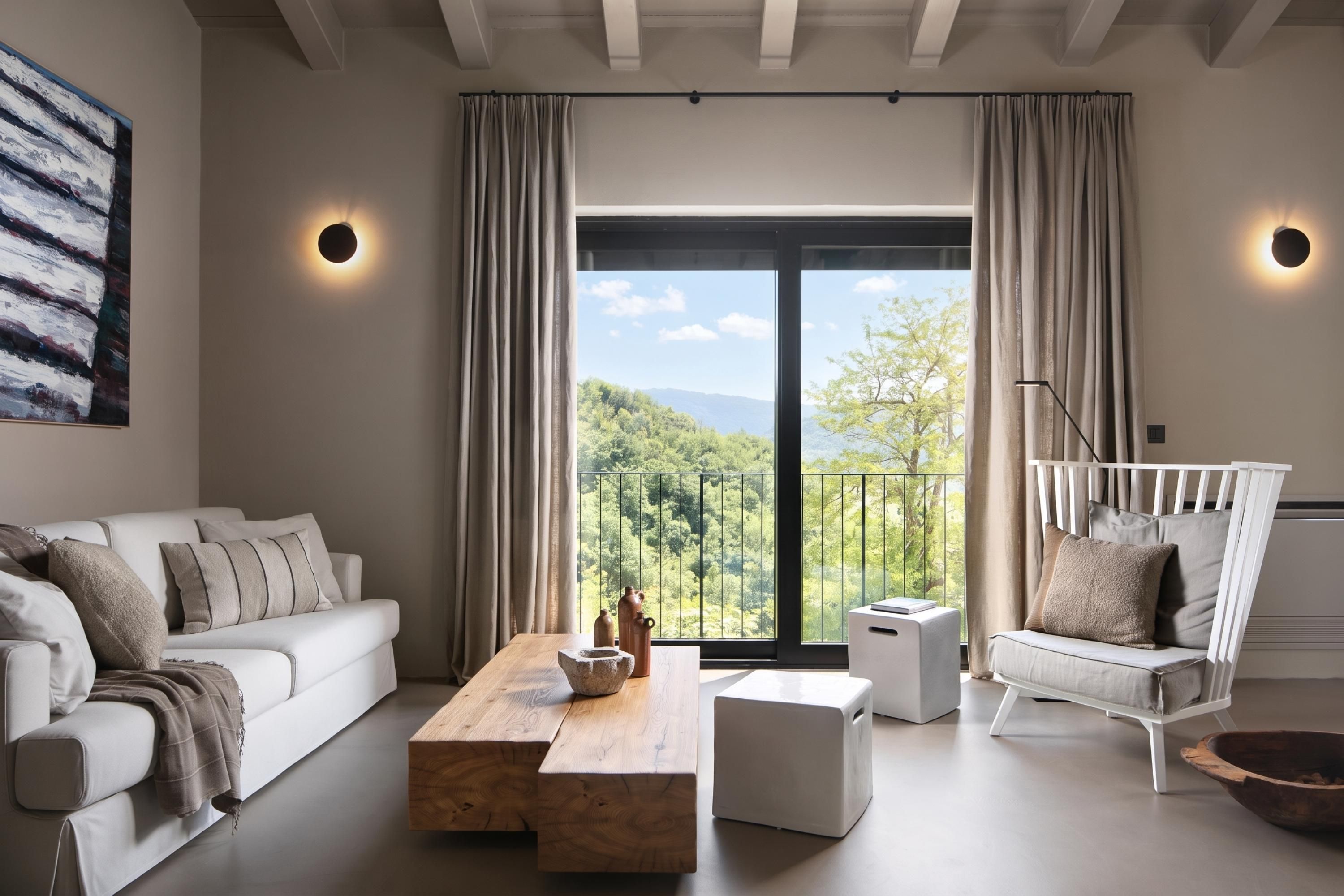 Un salon moderne avec un canapé blanc, une table basse en bois et un fauteuil blanc. Une grande fenêtre offre une vue sur une forêt et des montagnes.