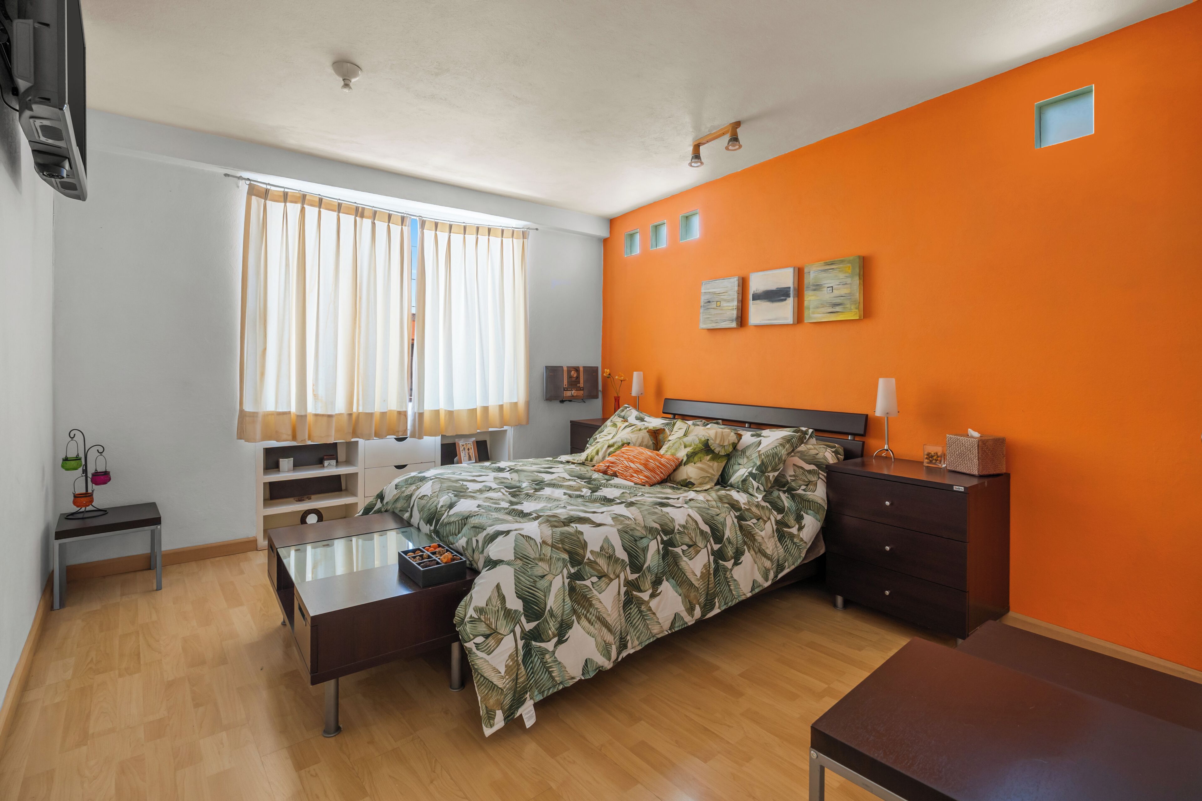 A bedroom with a bed, a bedside table, a dresser, a window with curtains, and a wall with three small pictures.
