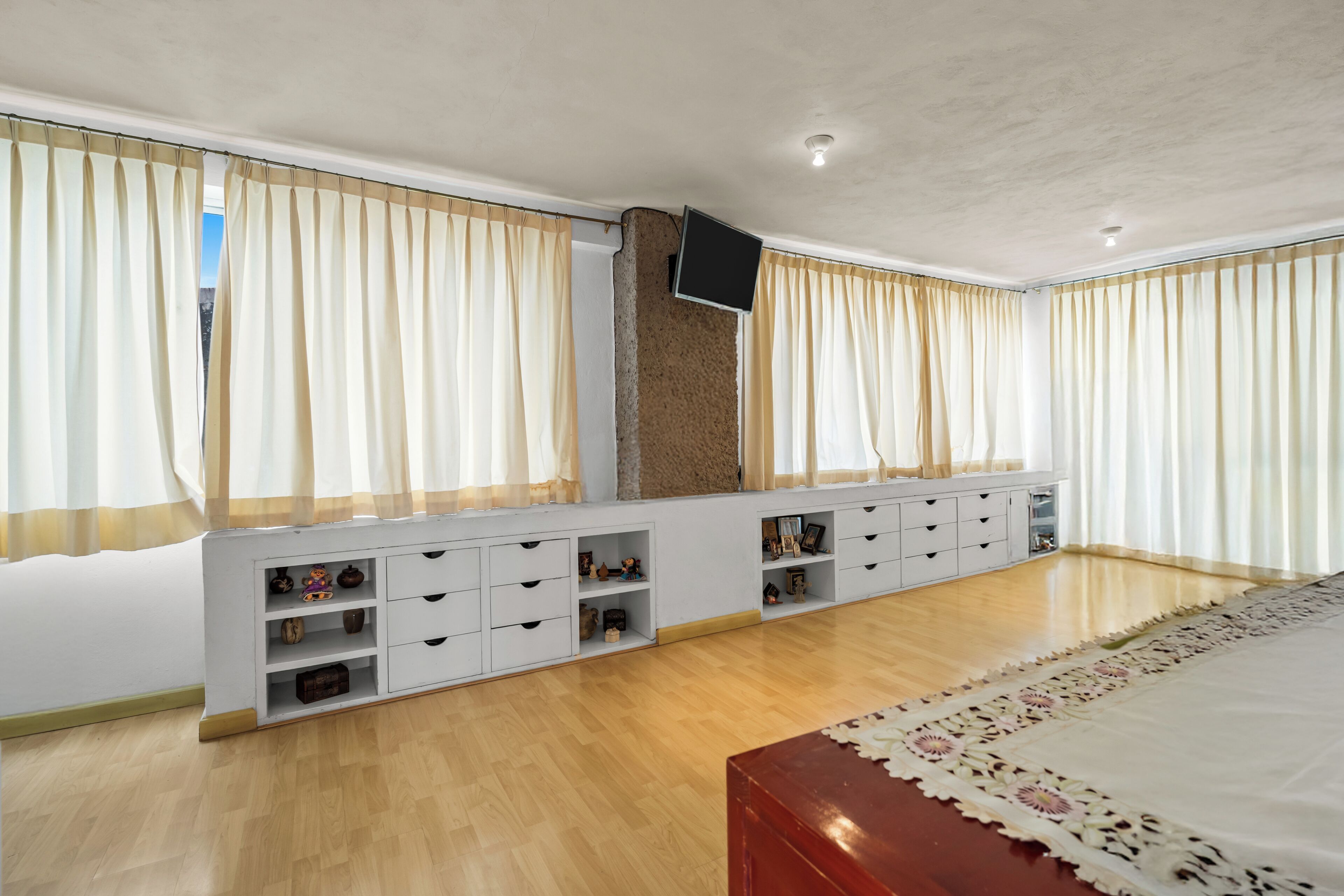A bedroom with a wooden floor, a bed with a floral-patterned bedspread, a TV mounted on the wall, and a built-in storage unit with multiple drawers.