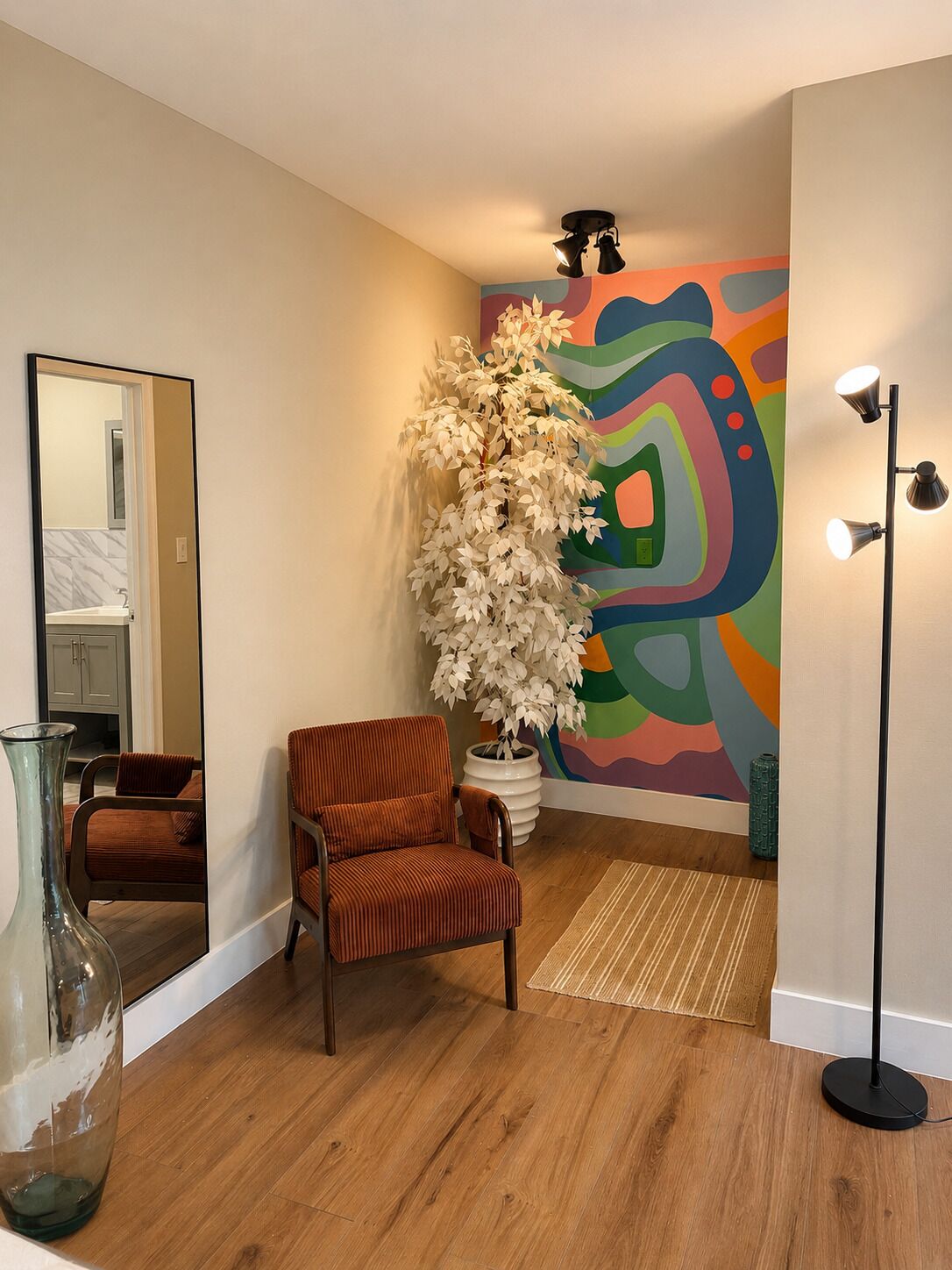 Une pièce avec un plancher en bois, une chaise, une grande plante, un miroir et une peinture murale abstraite aux couleurs vives.