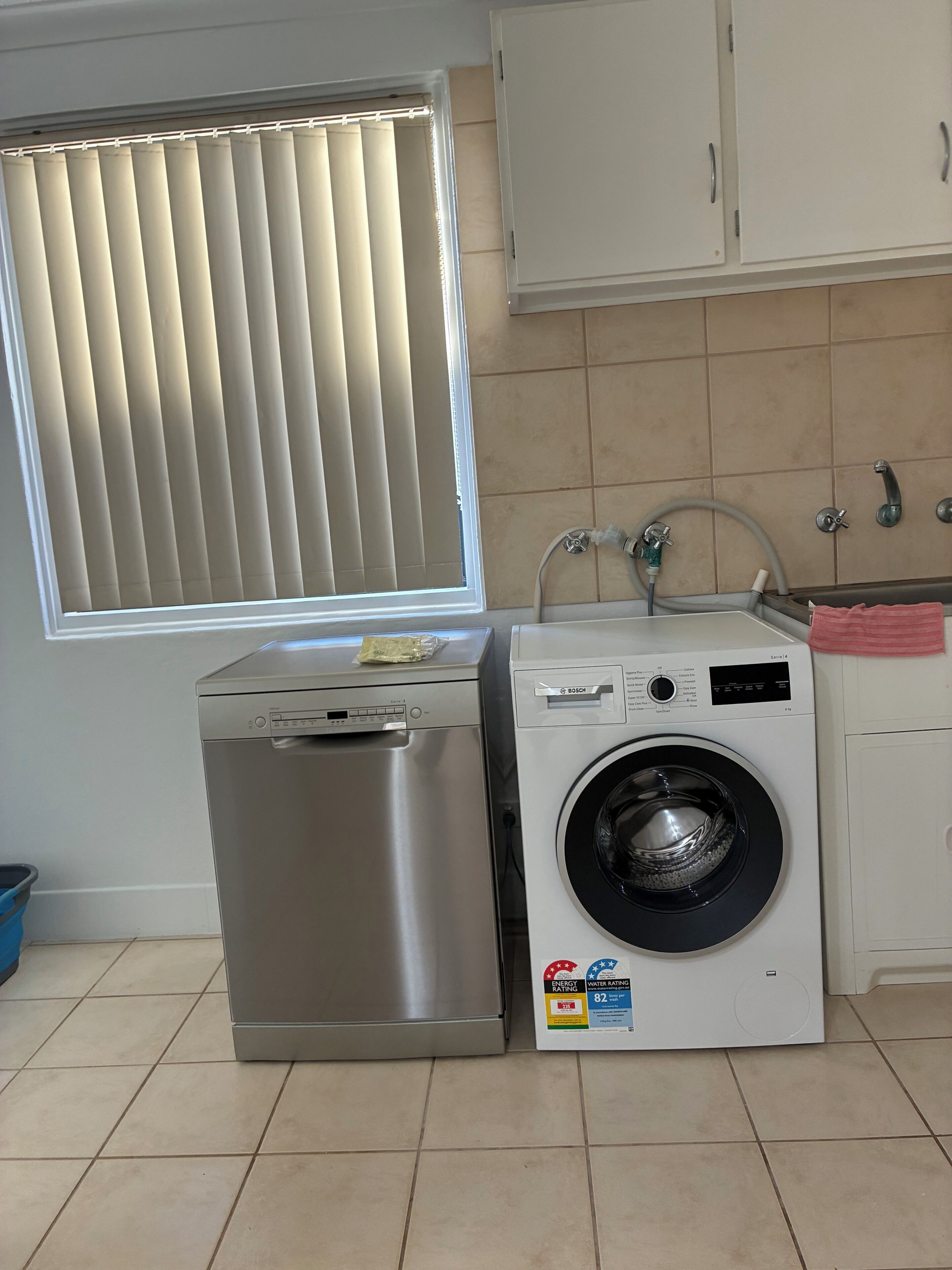 A compact kitchen with a washing machine, a stainless steel dishwasher, and a window with blinds.