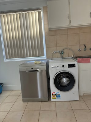 A compact kitchen with a washing machine, a stainless steel dishwasher, and a window with blinds.
