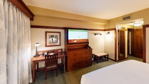 A hotel room with a bed, a desk with a lamp, a television, a chair, a small table, and a wardrobe.