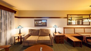 A hotel room with a sofa, a small table, a bench, a bedside table, a lamp, a mirror, and a framed picture on the wall.